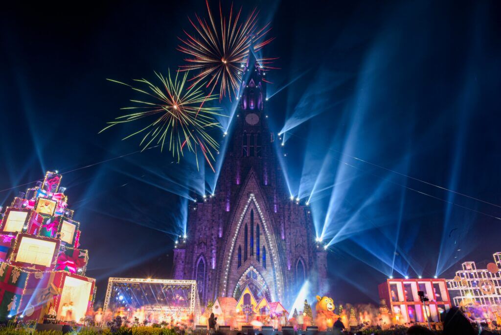 Imagem da Catedral de Pedra de Canela iluminada com várias luzes azuis e roxas, também fogos coloridos no céu. Também a Praça da Matriz decorada com vários enfeites de natal e com uma enorme árvore de presentes.