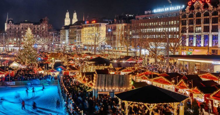 Imagem da cidade de Zurique de noite toda iluminada, do lado esquerdo uma grande árvore de natal e abaixo, neve.