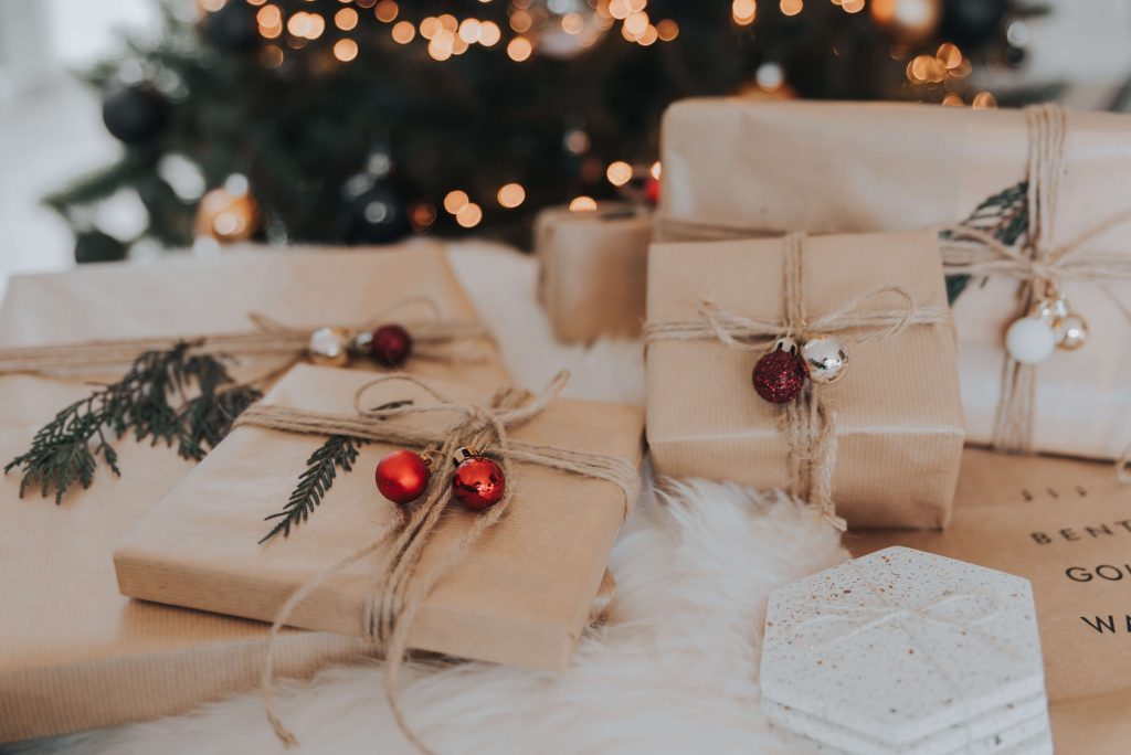 foto de vários presentes, embrulhadas com papel pardo e decoradas com bolas de natal. Ao fundo, de forma desfocada, há uma árvore de natal com luzes douradas.