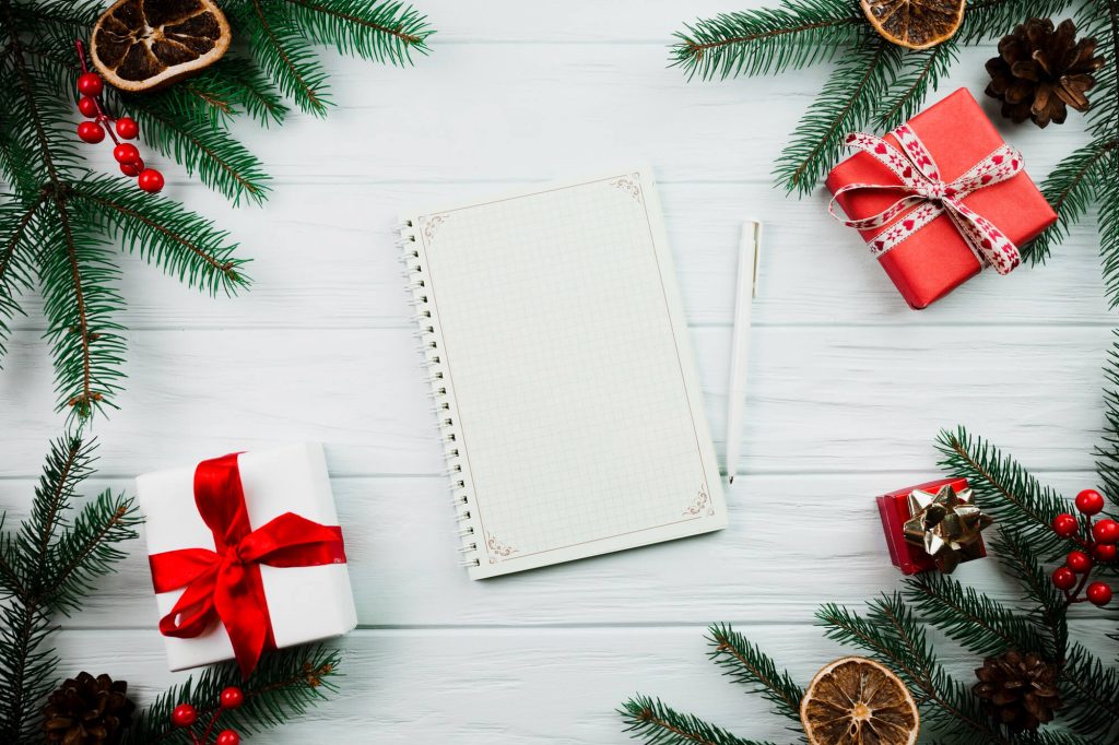 Imagem de um caderno em branco, uma caneta branca ao lado e em volta enfeites de natal verdes e vermelhos; e pequenos presentes com laços vermelhos.