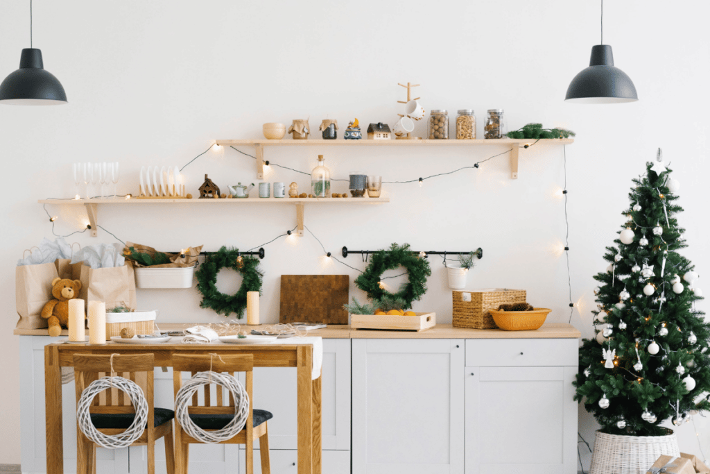 imagem de uma cozinha decorada com arranjos de natal, com armários brancos, com bancada e mesa de madeiras claras naturais, ao lado uma árvore de natal com enfeites brancos. 
