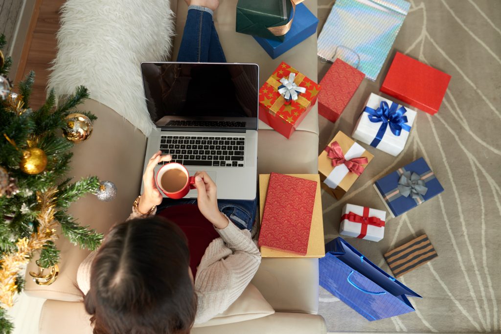 imagem tirada de cima, com uma mulher sentada em um sofá, tomando chocolate quente, olhando o laptop. Ao redor alguns presentes, cadernos e árvore de natal.