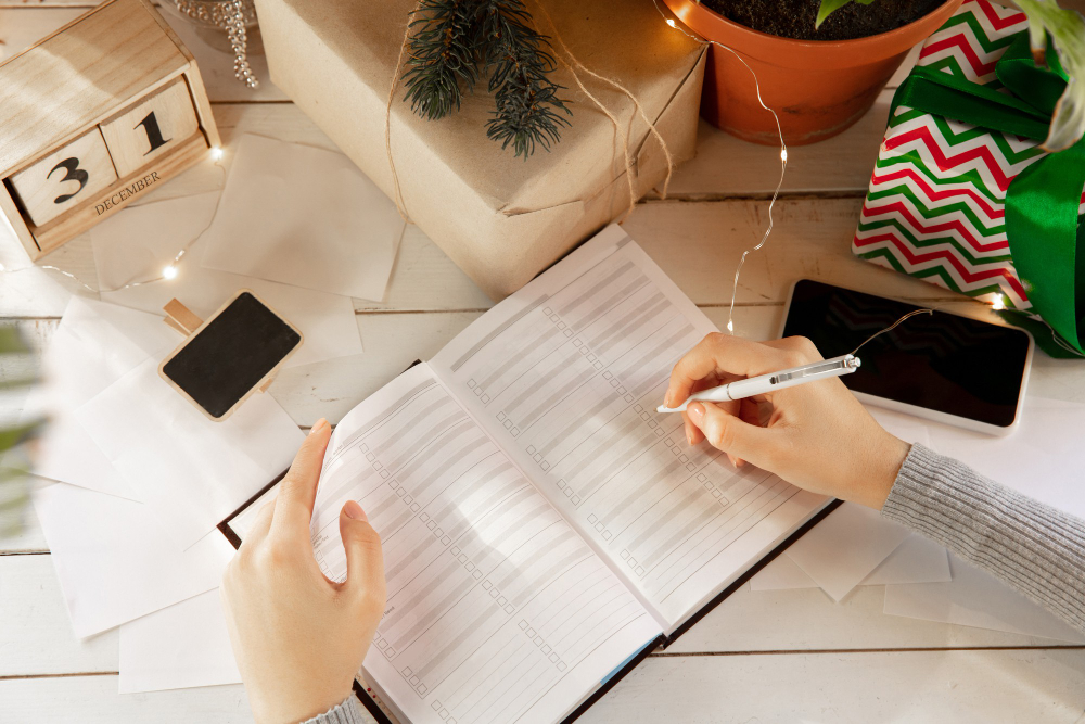 mãos femininas escrevendo em um caderno, ao redor vários itens de Natal. 